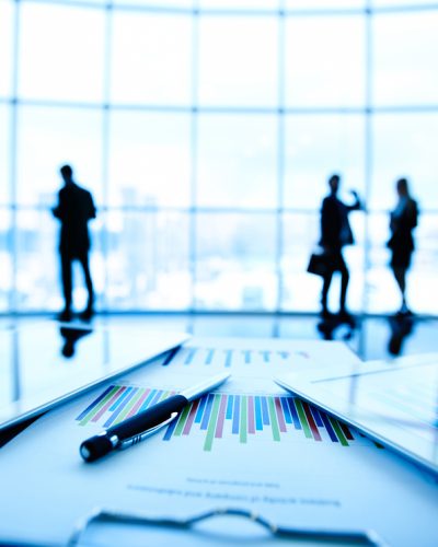 Technological devices and financial document with pen at workplace on background of three business partners communicating